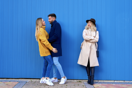A woman stands alone to the side of an embracing couple, considering them closely.